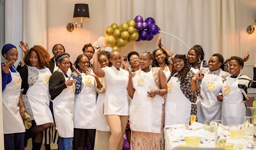 Neo and a group of students showing their finished cosmetic products, standing in front of a putple and gold balloon arch